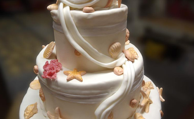 Torta di matrimonio ricoperta in pasta di zucchero con decori a tema marino, con conchiglie e drappi in pasta di zucchero